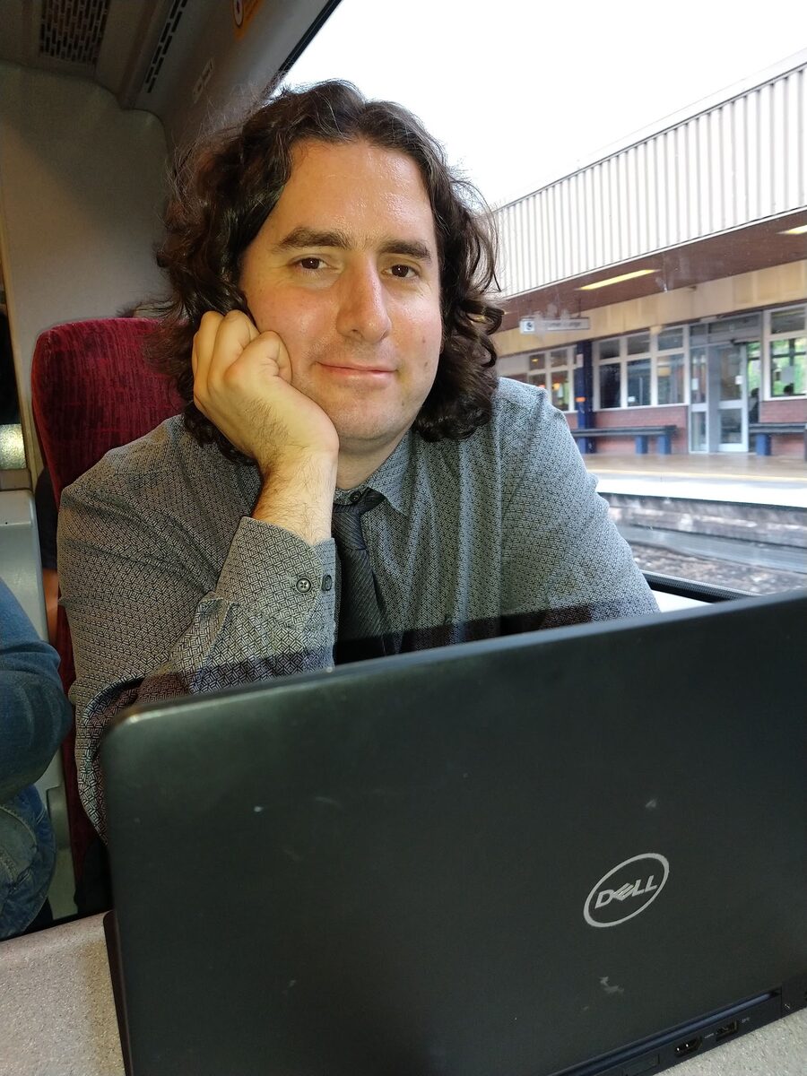 Zeth sitting at a laptop on a train, smiling at the camera